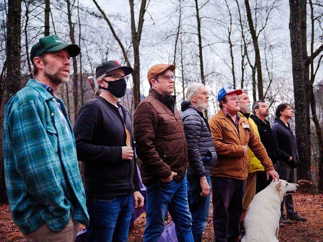 Eight people and a dog looking at something in the woods.