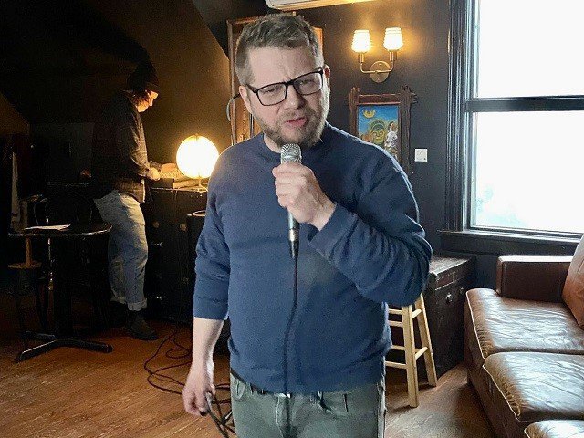 Nate Chappell, wearing dark-rimmed glasses and a blue shirt, holds a microphone.