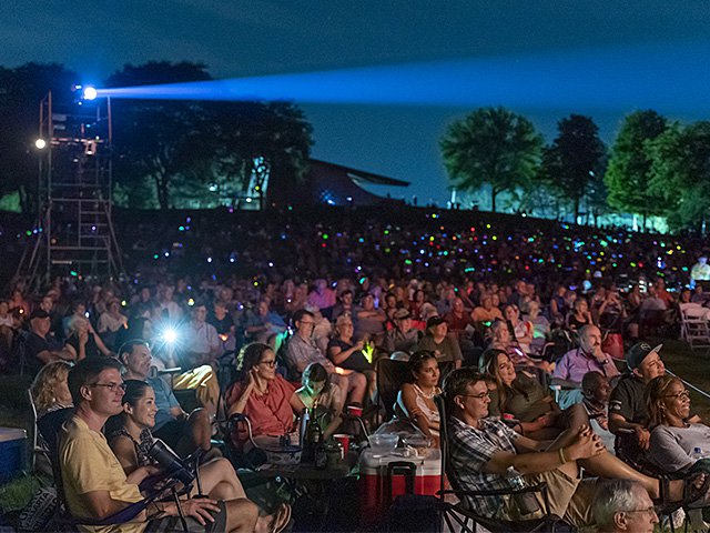 A crowded park audience watching opera at dusk.