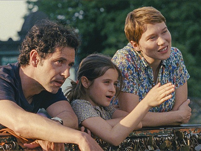 A man, a young girl of about six years of age and a teenager with short red hair lean on a railing and are looking at something off screen.