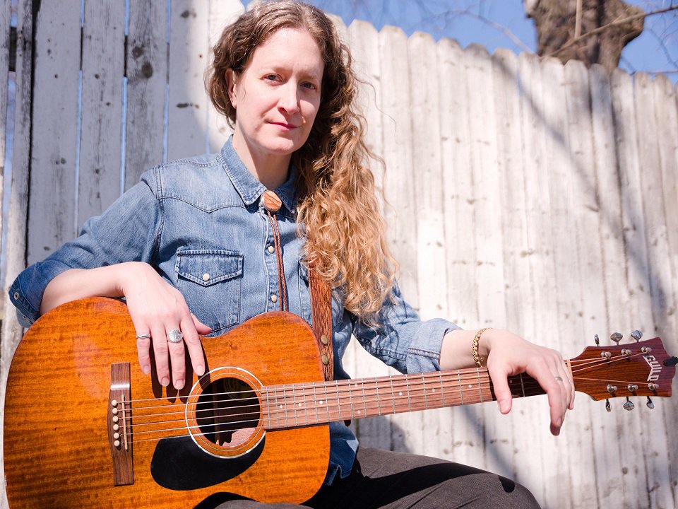 Marian Runk and guitar in front of a board fence.