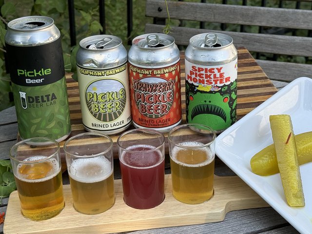 Cans of local pickles with a flight of them in glasses, and a couple of dill pickles.