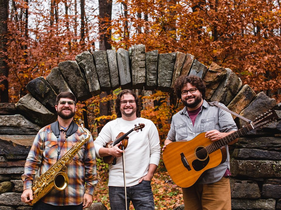 The three members of Faux Paws with instruments in front of a rock arch in the woods.