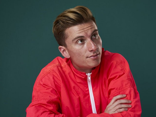 Comedian Trevor Wallace with a red jacket against a green background.