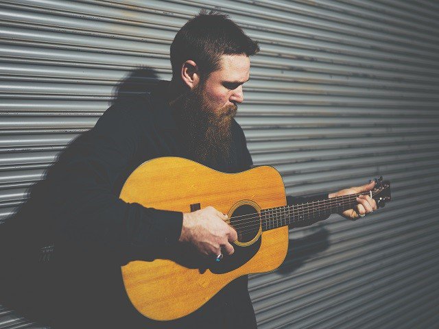 Andy Hughes with guitar in front of a corrugated steel wall.