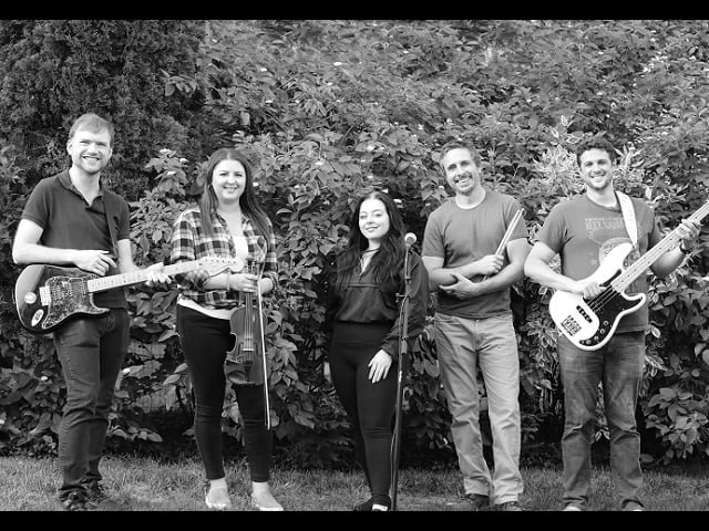 Five band members with instruments in front of a hedge.
