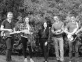 Five band members with instruments in front of a hedge.