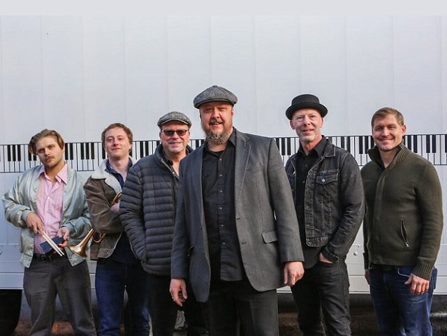 A six piece band in front of a wall.