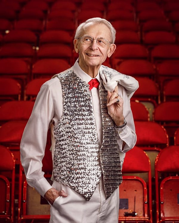 A man with a shiny silver vest stands in front of theater seating.