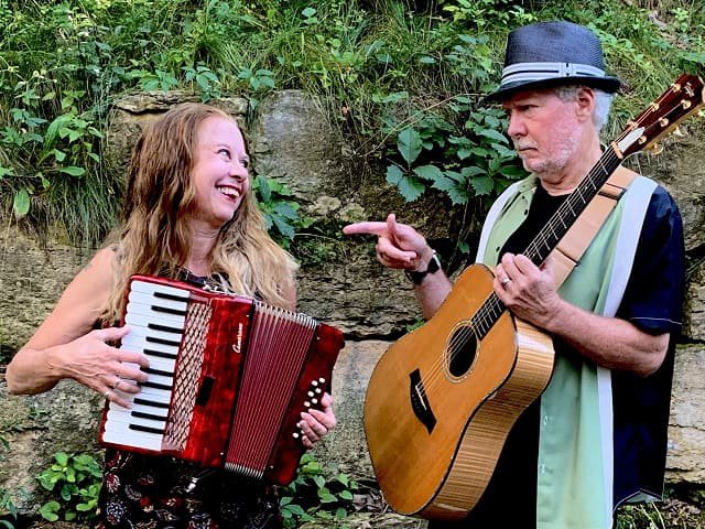 A man with a guitar points at a woman with an accordion.