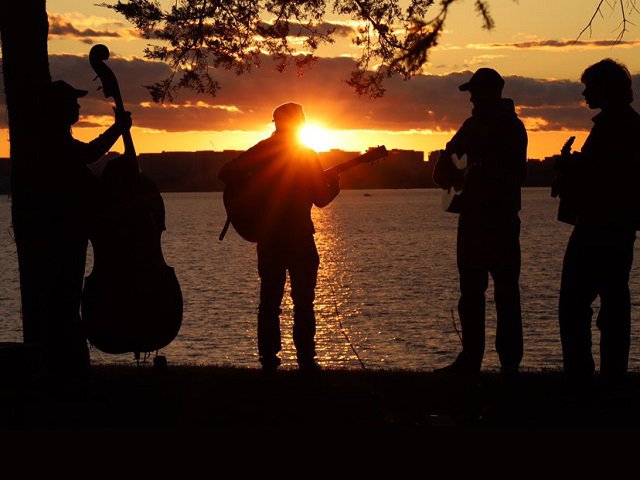 Four people with instruments in front of a lakeside sunset.