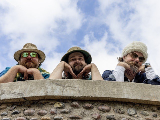 Three bearded individuals leaning on a stone wall.