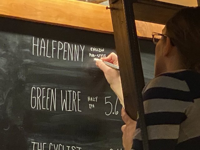 A brewery employee writes the name Halfpenny on a beer menu chalkboard.