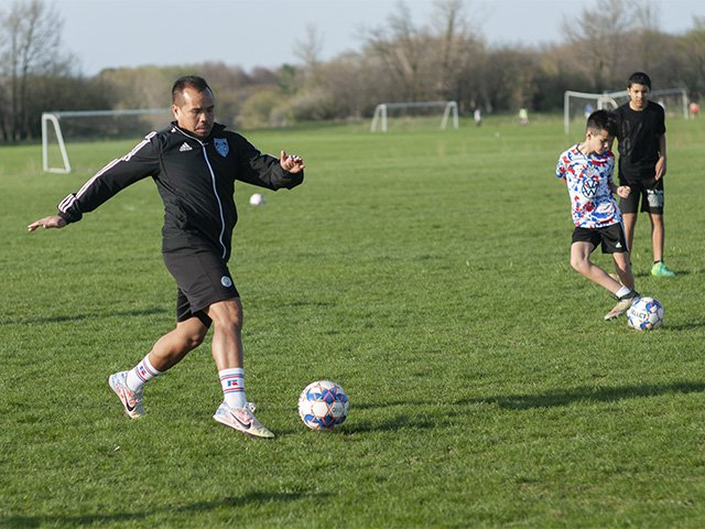 A man is about to kick a soccer ball while two younger children are also doing footwork with a soccer ball.