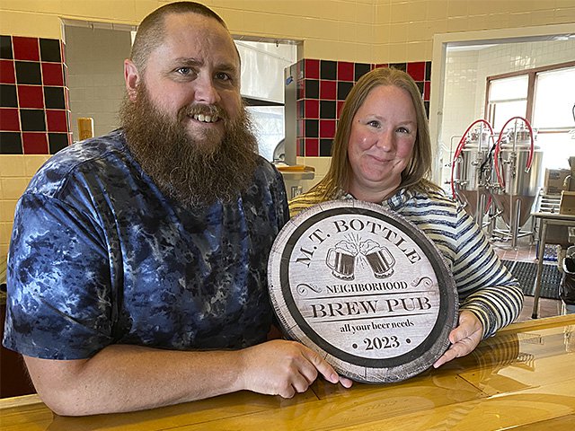 Mike and Heather Thompson are holding the sign for their new pub, in the new pub. Brewing equipment is in the background.