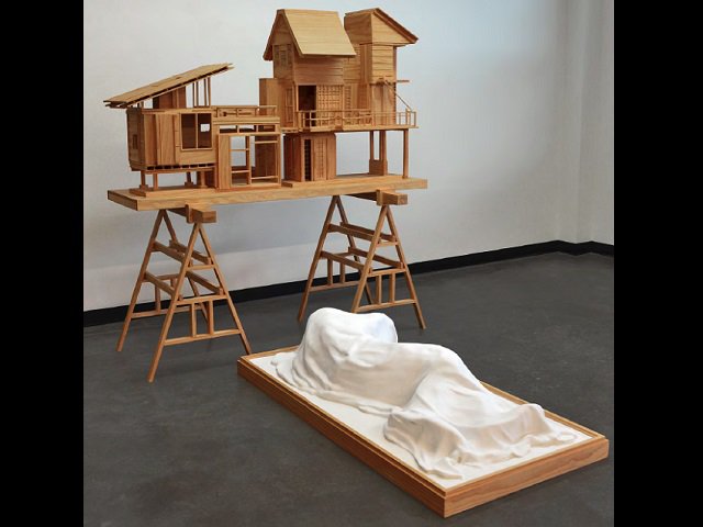 A model of a house on sawhorses with a figure under a sheet in front of it.