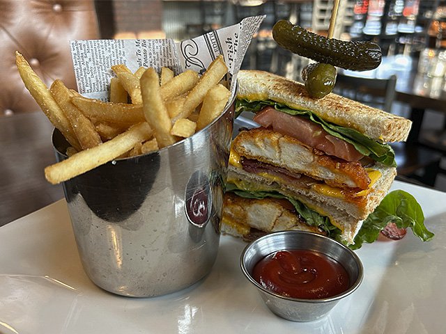 A silver container holding french fries and a highly stacked club sandwich topped with a two pickles in an almost sculptural arrangement.