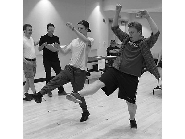 Two men in casual clothes are practicing a leg kick in a dance studio, their arms raised in the air.