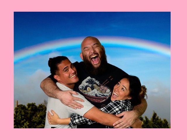 A large man is hugging a smaller man and a woman with sky and a rainbow in the background.