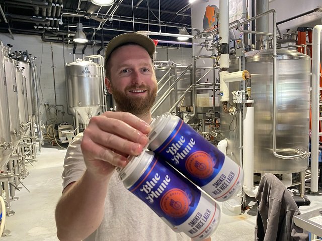 Clint Lohman in the brewhouse with cans of the beer Blue Plume.