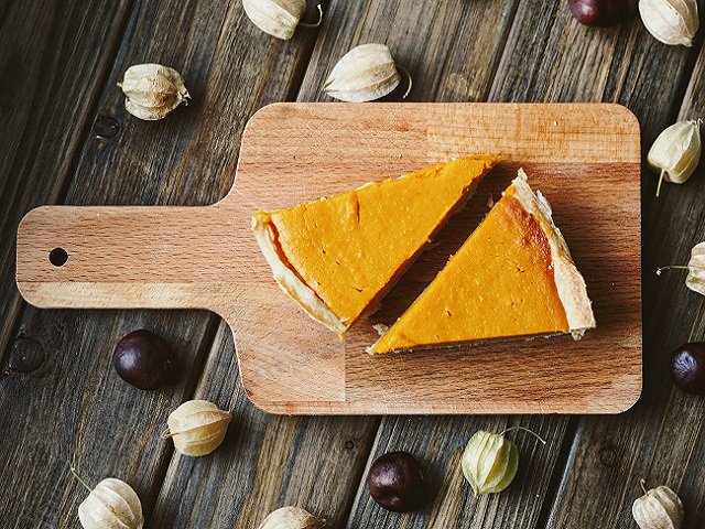 Two pieces of pumpkin pie on a wooden cutting board surrounded by nuts.