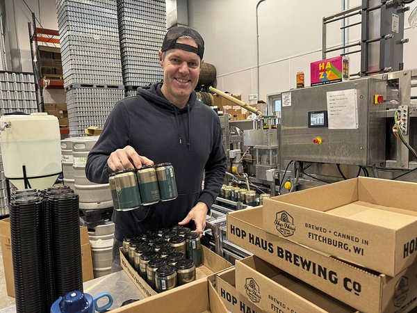 Phil Hoechst wearing a gray hoodie and holding a six pack in some sort of distribution area at the brewery.