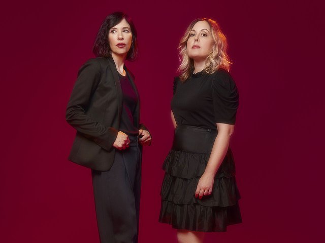 Carrie Brownstein wearing a gray suit and Corin Tucker wearing a black shirt and ruffled skirt.