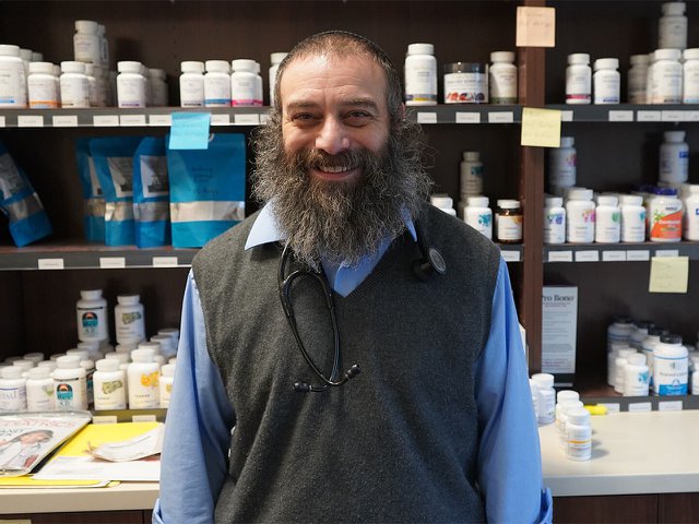 A bearded man with a stethoscope around his neck in front of a pharmacy-like shelf of pill bottles.