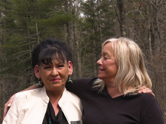 Two women, Vicky Harrison and Deborah Mejchar; Mejchar has one arm around Harrison's shoulder. They are standing among pine trees.