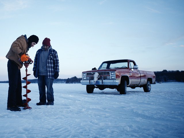 Two men dressed warmly stand on a frozen lake with an ice auger and with a red pickup truck in the background.
