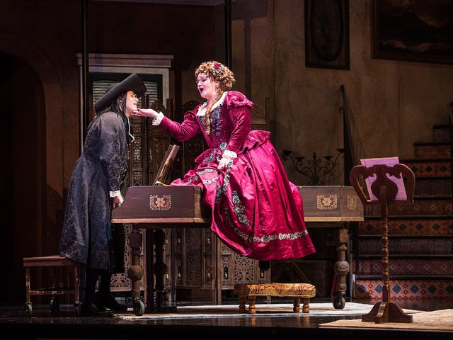 A woman, Rosina, sits on a piano as a man stands at the keyboard.