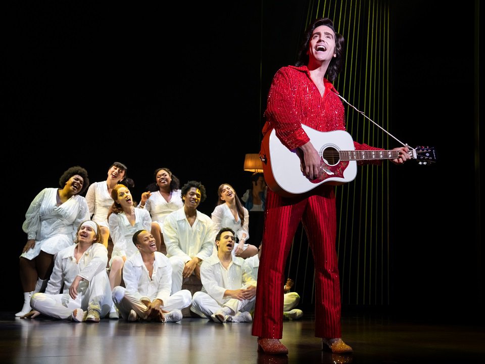 A person in a red suit with a white guitar in front of a choral group.